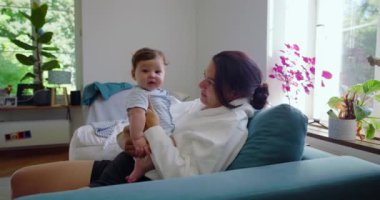 Anne, kanepede oturup gülümserken ve pencerenin doğal ışığıyla, sıcak bir atmosfer ve ev bitkileriyle çevrili rahat bir evde etkileşime girerken bebeği kucağına alıyor.