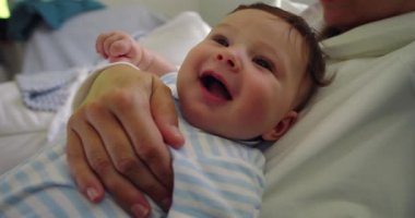 Bebek, annesinin göğsünde dinlenirken sevinçle gülüyor. Yumuşak doğal ışıkla sıcak bir yuva ortamında iç açıcı ve neşeli bir an yaratıyor.