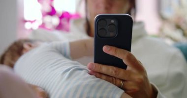 Anne, bebeğini kucağında tutarken telefonunu karıştırıyor. Bebek, kollarında huzur içinde, rahat ve aydınlık bir evde dinleniyor.