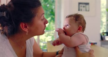 Anne bebeğini nazikçe kucağında tutuyor ve bir masada neşeli bir etkileşime giriyor. Bebek, aydınlık bir evde mutlu bir şekilde tepki veriyor.