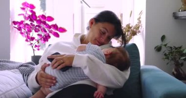 Anne bebeğini kollarında tutarken sıcak bir şekilde gülümsüyor. Yumuşak ışıkla sıcak bir ev ortamında şefkatli ve şefkatli bir an yaratıyor.