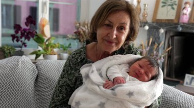 Büyükannem kanepede oturuyor, kundaklanmış yeni doğmuş bir bebeğe gülümsüyor, samimi bir yuva ortamının sevgi dolu bağını ve neşesini vurguluyor.