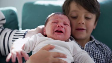 Küçük çocuk kucağında ağlayan yeni doğan kardeşi, sıcak bir ev ortamında kardeşler arasındaki duygusal karmaşıklığı ve hassas bağı vurguluyor.