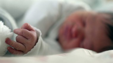 Yeni doğmuş bir bebeğin narin eline yakın çekim. Huzur içinde dinlenirken yumuşak beyaz bir battaniyeyi kavrayıp güzel bulanık arkaplanlı ilk yaşam anlarının masumiyetini ve hassasiyetini gösteriyor.