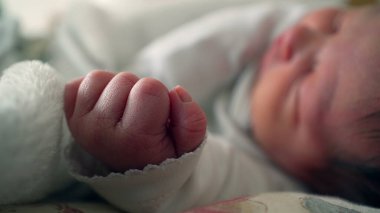 Yatakta huzur içinde uyuyan yeni doğmuş bir bebeğin elinin yakın çekimi. Küçük el, ilk yaşamın hassas ve hassas yönlerini ve yeni başlangıçları vurgulayan odak noktasıdır.