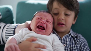 Küçük çocuk kucağında ağlayan yeni doğan kardeşi, sıcak bir ev ortamında kardeşler arasındaki duygusal karmaşıklığı ve hassas bağı vurguluyor.