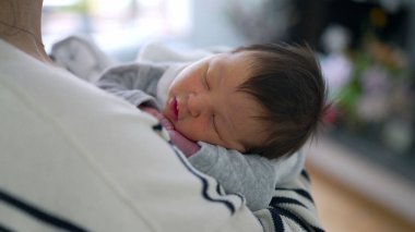 Anne omzunda huzur içinde uyuyan yeni doğmuş bir bebeğin yakın plan çekimi, anne ve çocuk arasındaki sıcaklığı, güvenliği ve derin bağları vurguluyor.