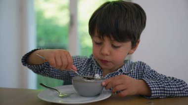 Küçük çocuk kaşıkla kahvaltıdan zevk alıyor. Konsantrasyon ve keyif gösteriyor. Mutfak ortamındaki ahşap bir masada oturuyor.