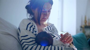 Anne sevecen bir şekilde kollarında yeni doğmuş bebeğe bakıyor. Huzurlu ve samimi bir yuvadaki şefkatli ve şefkatli bağı vurguluyor.