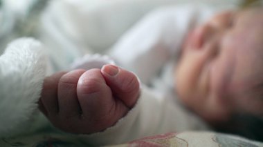 Bebek arka planda huzur içinde uyurken yeni doğmuş bir bebeğin elinin yakın çekimi. Erken yaşların şefkati ve narin doğası, bebekliğin sıcaklığını ve güvenliğini vurguluyor.