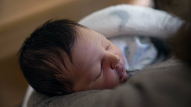 Annesinin kollarında uyuyan yeni doğmuş bir bebeğin yakın plan çekimi, erken yaşamın huzur ve güvenliğini gösteriyor. Erken yaşta verilen hassas bağ ve yetiştirme bakımı