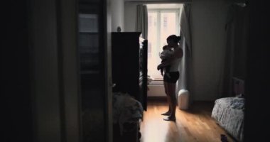 Anne pencerenin yanında dururken bebeğin kucağına usulca sarılıyor. Bağlantının ve aşkın samimi ve huzurlu anını paylaşıyor.