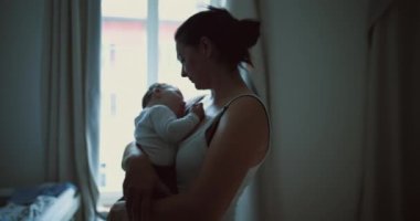 Anne pencerenin kenarında duruyor, bebeği kucaklıyor ve sevgi ve sevgiyle aşağı bakıyor, evde huzurlu ve samimi bir aile anı yakalıyor.