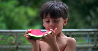 Çocuk bir dilim karpuz yemeye, balkonda meyvenin tadını çıkarmaya, huzurlu ve rahat bir ortamda anın tadını çıkarmaya odaklanmış.