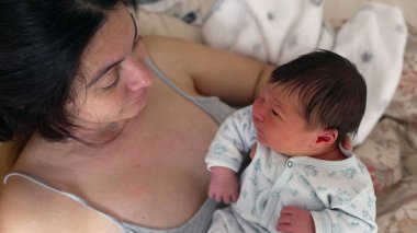 Anne, yeni doğmuş bebeğini nazikçe kucaklıyor. Bebek ağlıyor gibi görünüyor. Hayatının ilk günlerinde anne ve baba ile çocuk arasındaki bağın erken yaşta ortaya çıktığı duygusal bir an.