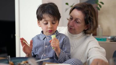 Küçük çocuk yemek yerken büyükannesinin kucağında oturuyor, yemeğine odaklanıyor. Büyükannem onunla bir diyalog içinde, şefkatli, şefkatli bir aile etkileşimi gösteriyor.