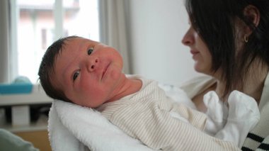 Anne yeni doğmuş bebeği kucağına alıyor, bebeğin gözleri tamamen açık sakin bir ifadeyle, anne ve çocuk arasındaki erken bağ, hassas ve samimi bir bağ oluşturuyor.