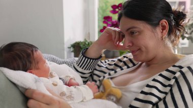 Anne kanepeye yaslanmış, yeni doğmuş bebeğine bakarken yavaşça gülümsüyor, rahat yastıklar ve canlı bitkilerle çevrili, huzurlu bir aile anı.