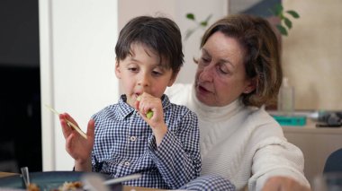 Küçük çocuk yemek yerken büyükannesinin kucağında oturuyor, yemeğine odaklanıyor. Büyükannem onunla bir diyalog içinde, şefkatli, şefkatli bir aile etkileşimi gösteriyor.