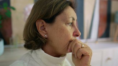 Derin düşüncelere dalmış, eli çenesinde dinlenen yaşlı bir kadın, derin bir sessizlik sırasında derin bir anda yakalandı evde, yumuşak doğal ışık onun düşünceli yüz ifadesini aydınlatıyor.