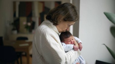 Yaşlı bir kadın yeni doğmuş bir bebeği nazikçe kucaklıyor, yumuşak ışıklı bir odada pencerenin yanında duruyor, bebeğe sakin ve şefkatli bir ifadeyle bakarken ona bakıyor.