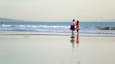 Sakin bir günde kıyı şeridi boyunca yürüyen iki insan, ıslak kumda yansımaları görünür, arka planda hafif dalgalarla huzurlu ve sakin bir plaj sahnesi yakalıyorlar.
