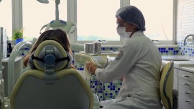 Diş hekimi, diş hekimliği koltuğunda oturan bir hastaya bakıyor. Modern bir klinikte, temiz ve parlak bir çevreye sahip diş tedavisi ve inceleme ekipmanlarını tartışıyor.