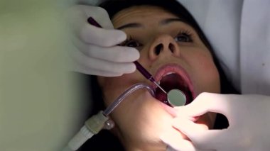 Diş hekimi hassas aletlerle çalıştığı için hastaların ağzı sonuna kadar açık. Parlak ışık doğrudan dişlerin üzerine parlıyor, dişçinin dikkatli ve profesyonel çalışmalarını vurguluyor.