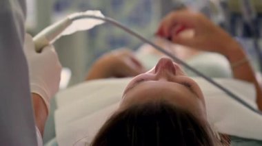 Hasta, dişçi koltuğunda uzanıyor. Yüzüne yakın çekim. Dişçi, iyi aydınlatılmış, temiz bir klinikte bir prosedüre hazırlanıyor.