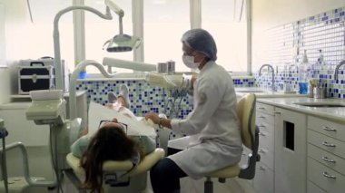 Diş hekimi, modern, temiz bir klinik ortamında dişçi koltuğunda yatan bir hastaya bakarken diş lambası ve aletler hazırlıyor.