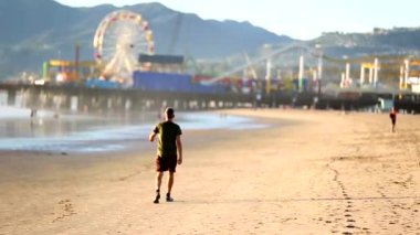 Kumsalda eğlence parkına doğru yürüyen bir adam, arka planda uzak dönme dolap, kumdaki ayak izleri, günlük gezinti, boş zaman, şehrin ufuk çizgisi