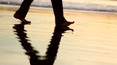 Çıplak ayaklı biri ıslak kumsalda yürüyor, gün batımında suda yansıyor, sakin dalgalarla sakin bir sahne, sakin bir an, rahatlama ve farkındalık, sahil yürüyüşü, huzurlu bir yalnızlık.
