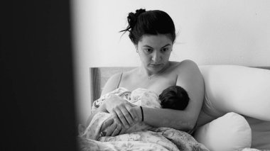 Anne yeni doğmuş bebeğini yatakta otururken emziriyor. Anneliğin ilk dönemlerinde anne ve çocuk arasında paylaşılan samimi bağın duygusal ve huzurlu tasviri, tek renk.