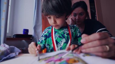 Genç bir çocuk, annesi dikkatlice izlerken bir çizim faaliyetinde bulunuyor. Ev ortamında sessiz ve odaklanmış bir aile anını paylaşıyor.