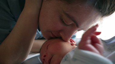 Anne yeni doğan bebeği alnından nazikçe öper, anne ve baba ile çocuk arasındaki sevgi ve bağın hassas bir anını gösterir. Erken ebeveynlikteki derin bağ ve şefkat