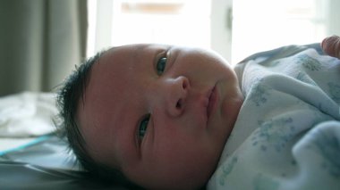 Yeni doğmuş bir bebeğin yatakta uzanmış, tamamen uyanık ve dünyayı merakla izleyen görüntüsü. Erken yaşların masumiyeti ve mucizesi huzurlu bir anda