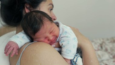 Anne, yeni doğmuş bebeğini omzunun üstünde tutarak bir anlık sakinlik ve bağ gösteriyor. Erken yaşta anne ve çocuk arasında paylaşılan derin bağ ve rahatlık.