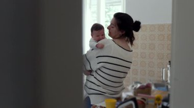 Anne sevgi dolu bir şekilde ağlayan bebeğini kucaklıyor ve öpüyor. Sıcak bir ev mutfağında samimi bir anda onu rahatlatıyor. Sıcaklık ve ebeveyn bakımı gösteriyor.
