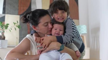 Anne ve büyük kardeş, kapalı bir aile ortamında bebeğe neşeyle sarılıyorlar. Anne, bebek ve kardeş arasındaki sevgi ve bağ sıcak ve sevgi dolu bir bağı paylaşıyor.