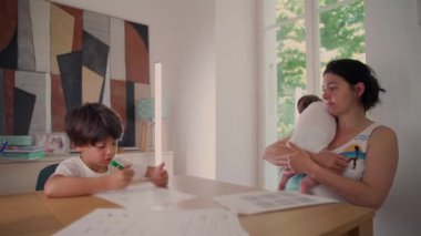 Anne arka planda bir bebeği tutarken çocuk masayı odak ile boyuyor, ev ortamında huzurlu bir aile anı yaratıcılık ve bakım kaynaştırıyor.