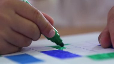 Yeşil kalemle kareleri doldururken konsantre olan çocuk, ev ortamında sanatsal bir aktivite sırasında dikkatini ve dikkatini çekiyor.
