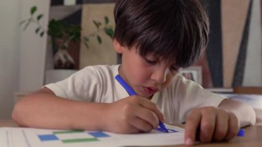 Bir sayfayı işaretleyici ile boyayarak konsantre olan genç çocuk, ev ortamında bir masada yaratıcı bir işle meşgul, odaklanmayı, öğrenmeyi ve çocukluk yaratıcılığını yansıtıyor.