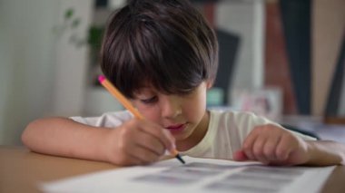 Kalemle çizim yapan genç bir çocuğun yakın plan çekimi, ev ortamındaki bir masada sanatsal çalışmalarına yoğunlaşması, çocukluk yaratıcılığı, öğrenme ve düşünceli bir odaklanma.