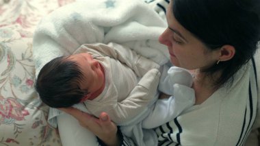 Anne yeni doğmuş bebeği yumuşak bir battaniyeye sarmış, sıcak ve huzurlu bir ev ortamında sevgi dolu, hassas bir bağ anına bakıyor, yukarıdan yakın çekim, sıcak ve samimi.