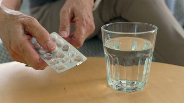 Elleriniz su toplayan bir bardaktan alınan hapı bastırıyor, bir ev ortamında ilaç ve sağlık hizmetlerini ön plana çıkarıyor.