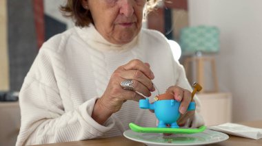 Yumurtayı çıkarmak için kaşık kullanan, rafadan yumurta yiyen yaşlı bir kadının yakın çekimi. Basit kahvaltı seti, sessiz ve rutin bir anın altını çizer.