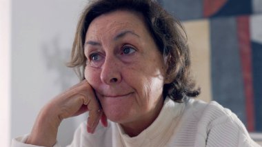 Yaşlı bir kadın masada oturuyor, kafası meşgul ve düşünceli bir ifadeyle elini uzatıyor. Sessiz bir ev ortamında tefekkür ve içgözlem anı