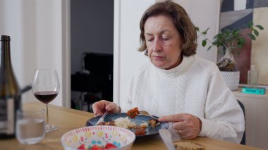 Evde yemek yiyen, çatalla büyük bir lokma yiyen yaşlı bir kadın. Akşam yemeğinin keyfini çıkartırken yakın plan, pilav ve bir kadeh şarap görünür. Ev yemeği, aile yemeği.