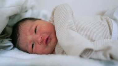 Yeni doğmuş bebek yatakta yatıyor, uyanık ve ince yüz ifadeleri takınıyor. Bebek sakin ama meraklı görünüyor. Erken yaşların huzurlu ve narin anı.