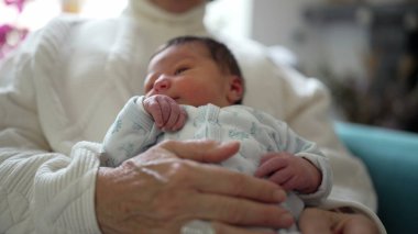 Yeni doğmuş bebek, büyük ebeveynlerinin elinde tutularak dünyaya merakla bakıyor. Farklı nesiller arasında paylaşılan değerli bağı ve sevgiyi vurguluyor.